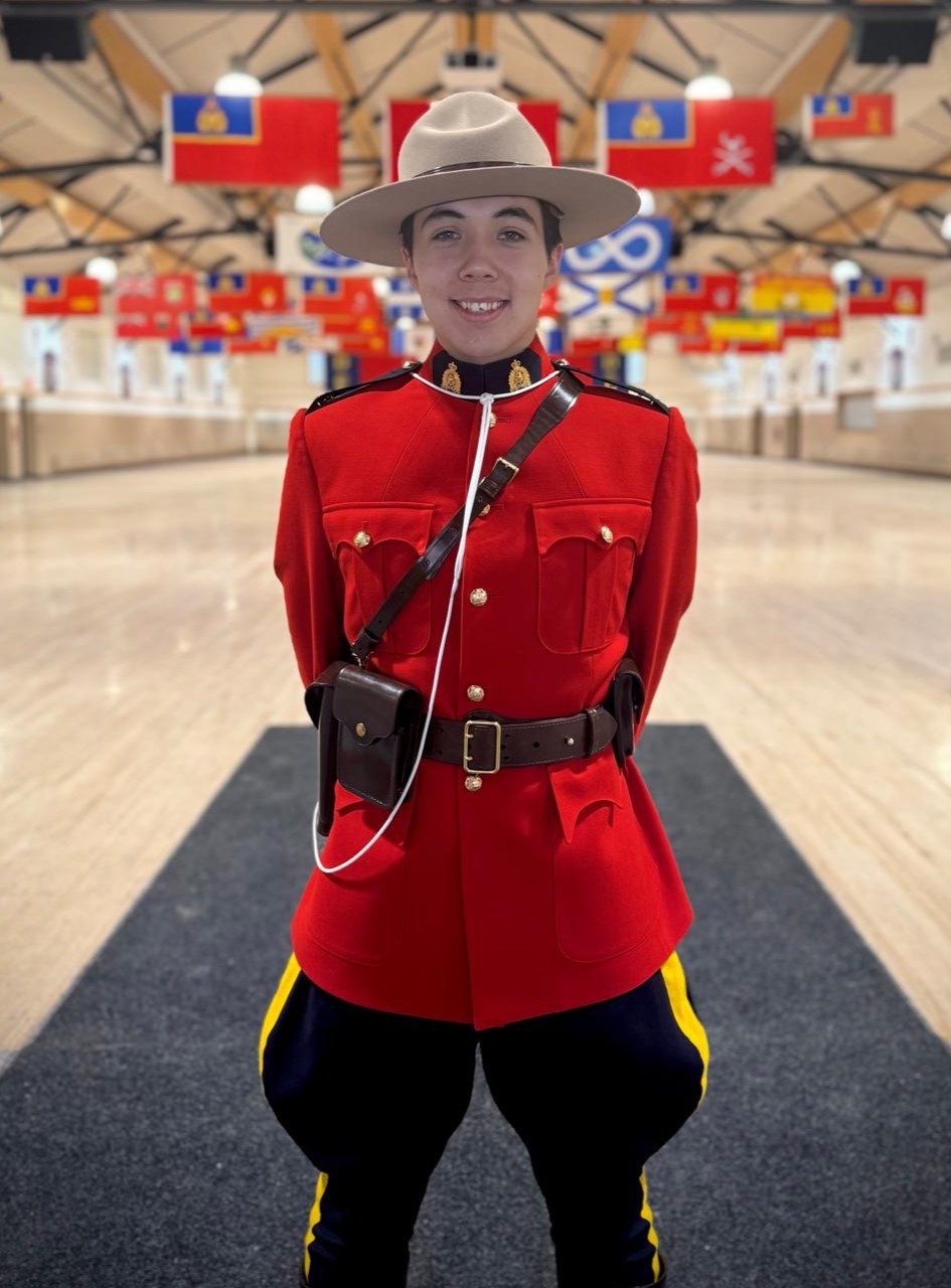 Male officer in Red Serge stands in a gym with Canada’s provincial flags hanging from the ceiling in the distance behind him.