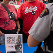 Two people stand across from a police officer in uniform. An 'Apply now' poster is displayed on the table between them.