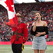 RCMP officer holding Canadian flag next to singer holding microphone standing on football field in front of stadium crowd.