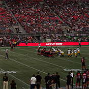 Footballers scrum on field - bright digital ads display RCMP recruitment messages around the stadium.