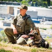 An RCMP officer in a tactical uniform crouches next to a German Shepherd dog on top of a hill.
