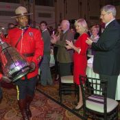Two RCMP officers, one male one female, in Red Serge carry the CFL grey cup in a room filled with tables and crowds of formally-dressed people watching.