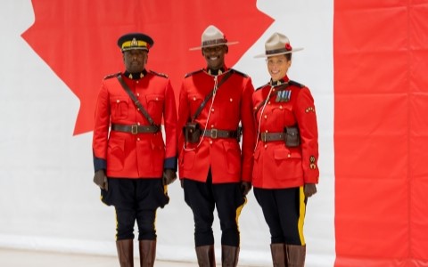 Two male and one woman officer wearing red serge smile for a photo in front of a wall mural of the Canadian flag.