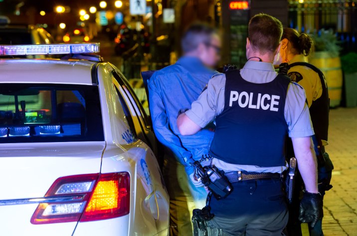 RCMP officers standing beside a marked police vehicle during a nighttime roadside interaction, with one individual positioned near the rear of the vehicle. Streetlights and urban surroundings are visible in the background.