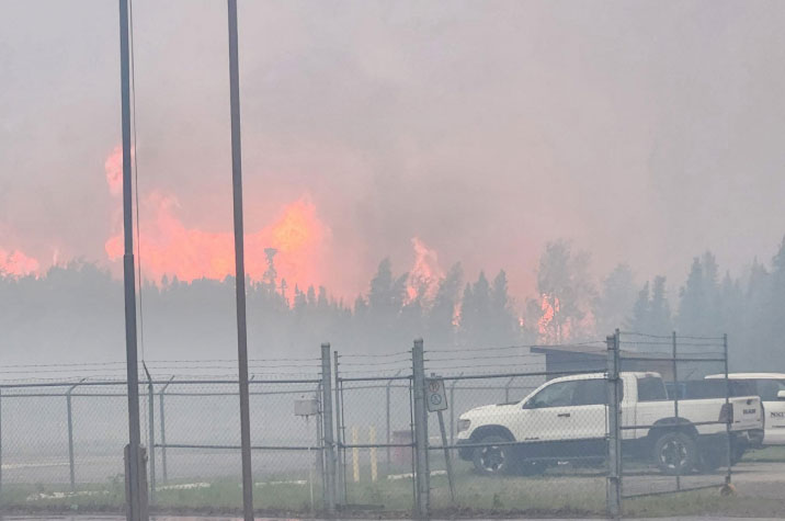 Au loin, un boisé est la proie de flammes rougeoyantes dont se dégage un panache de fumée gris foncé. En deuxième plan, deux véhicules blancs se trouvent dans une zone clôturée.