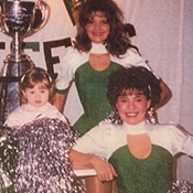 Two women cheerleaders pose for the camera with pom-poms. A young child sits in a giant grey cup between them. 