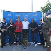 A group of male RCMP officers, in and out of uniform, pose in front of a big blue background with the words 'GRC/RCMP'. A German Shepherd dog sits in front of them.