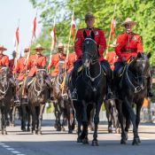 Two Mounties lead an escort of mounted riders carrying flagpoles.