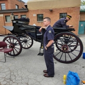 A man in blue coveralls leans stands next to a horse carriage. Another sits inside polishing the exterior. Image by Patricia Vasylchuk, RCMP.